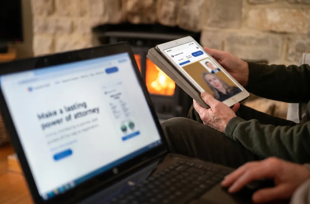 Someone sitting by a wood burner with a laptop on their knee, holding a tablet that shows a recorded video call in the Capacity Vault app.